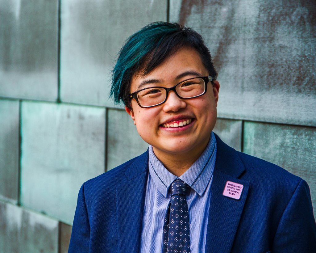Photo: Lydia smiles and tilts their head slightly to the side, looking confidently at the camera. They are a young-ish East Asian person with a streak of teal in their short black hair, wearing glasses, a cobalt blue jacket and navy tie, with a blue copper wall behind them. Photo by Sarah Tundermann